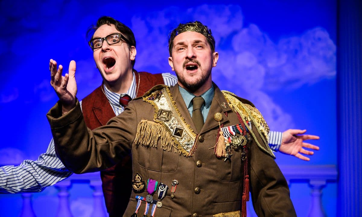 Two actors singing, one dressed as a king in front of the other wearing glasses and a waistcoat